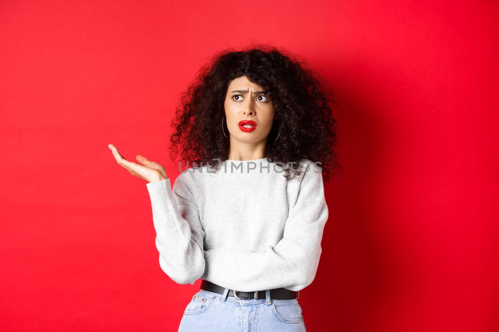 Annoyed and puzzled young woman with curly hair, raising hands up and looking aside, cant understand something strange, standing on red background.