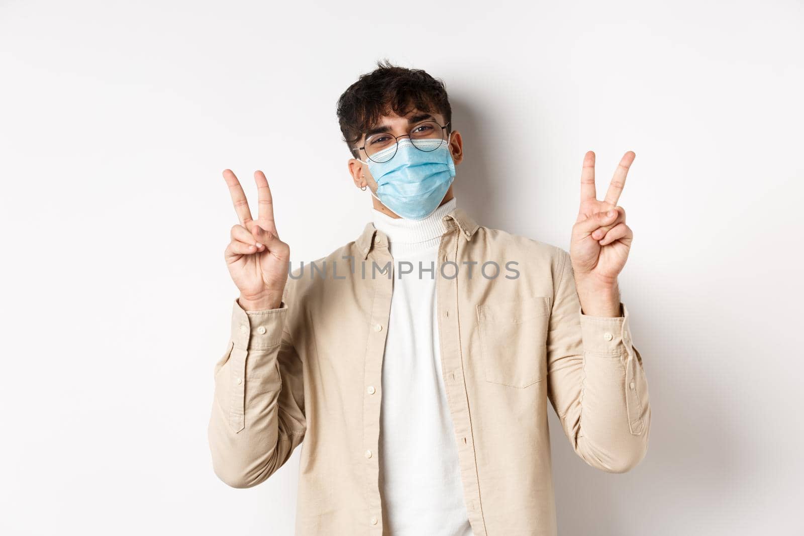 Covid-19, health and real people concept. Cute happy guy in medical mask staying positive, showing peace signs and smiling, standing on white background.