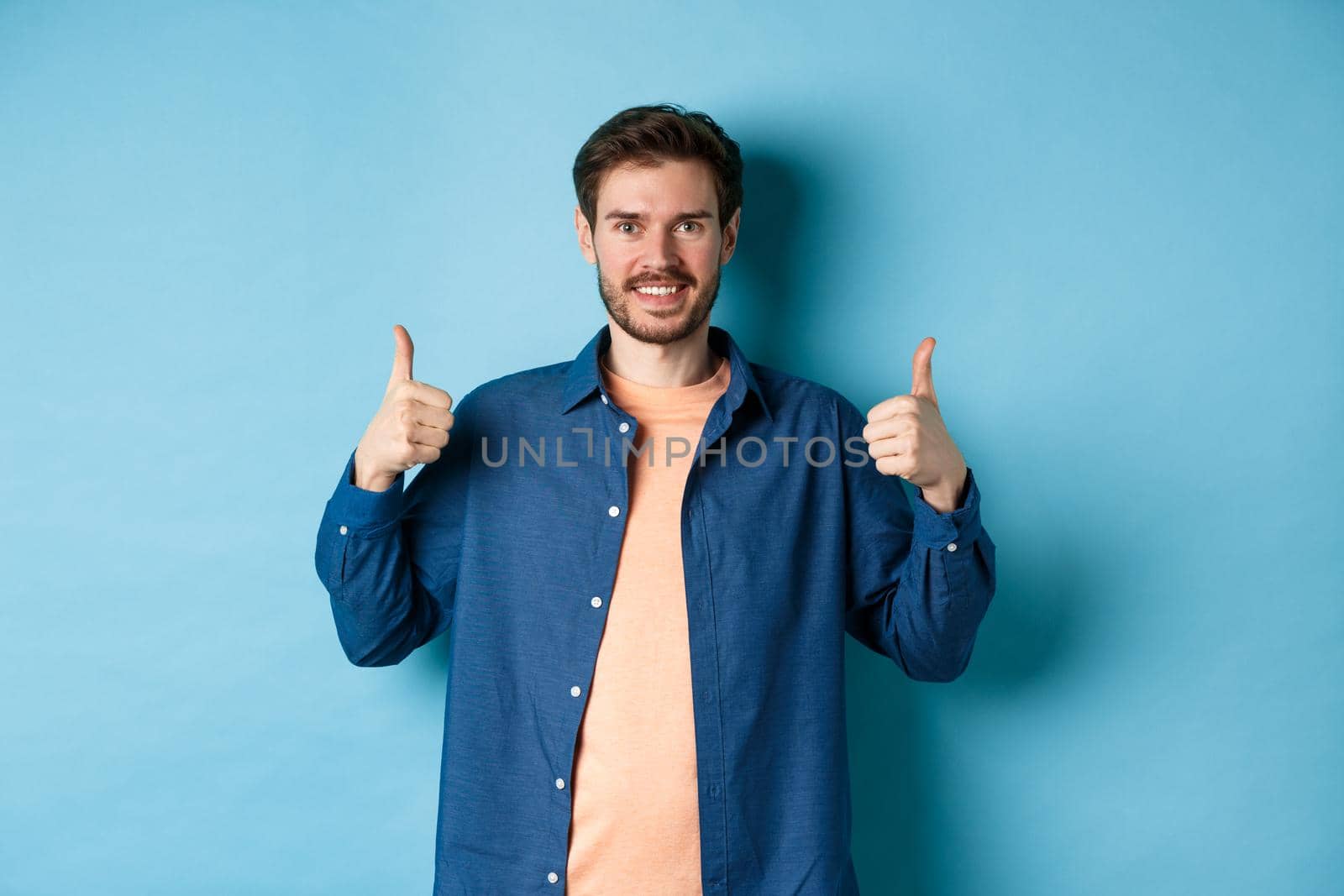 Handsome smiling man being supportive, showing thumbs up in approval, praise good work, standing happy on blue background.