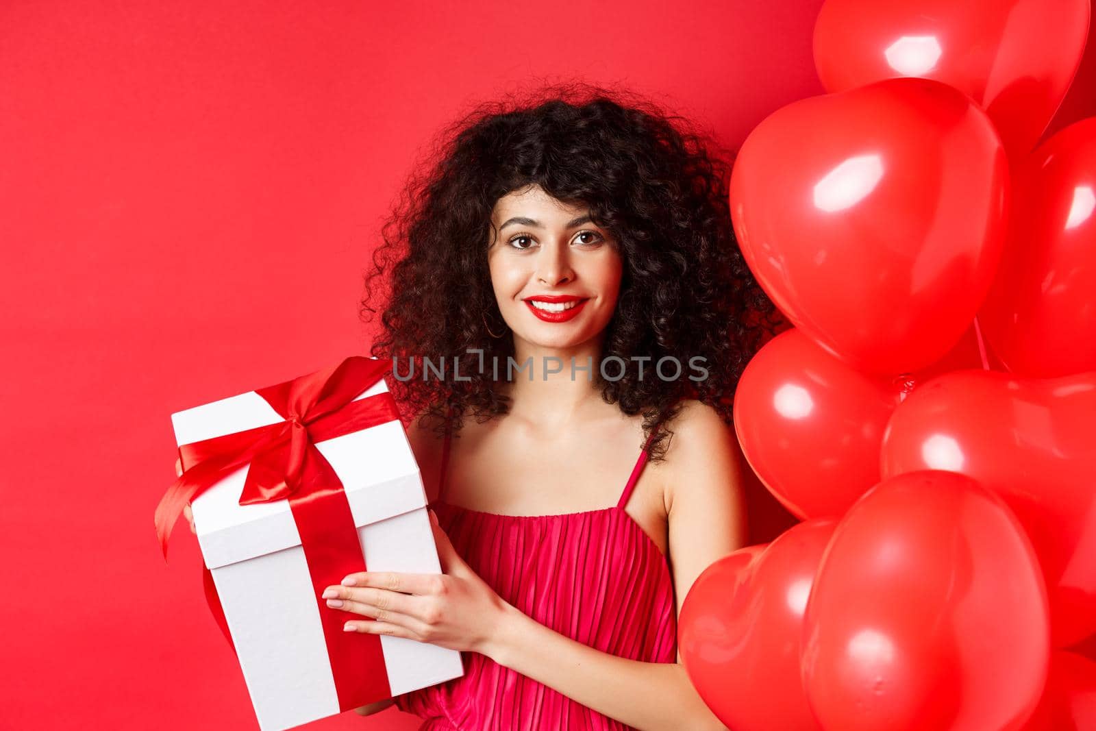 Holidays and celebration. Beautiful woman with curly hair, standing near heart balloons, holding gift box and smiling happy, white background.