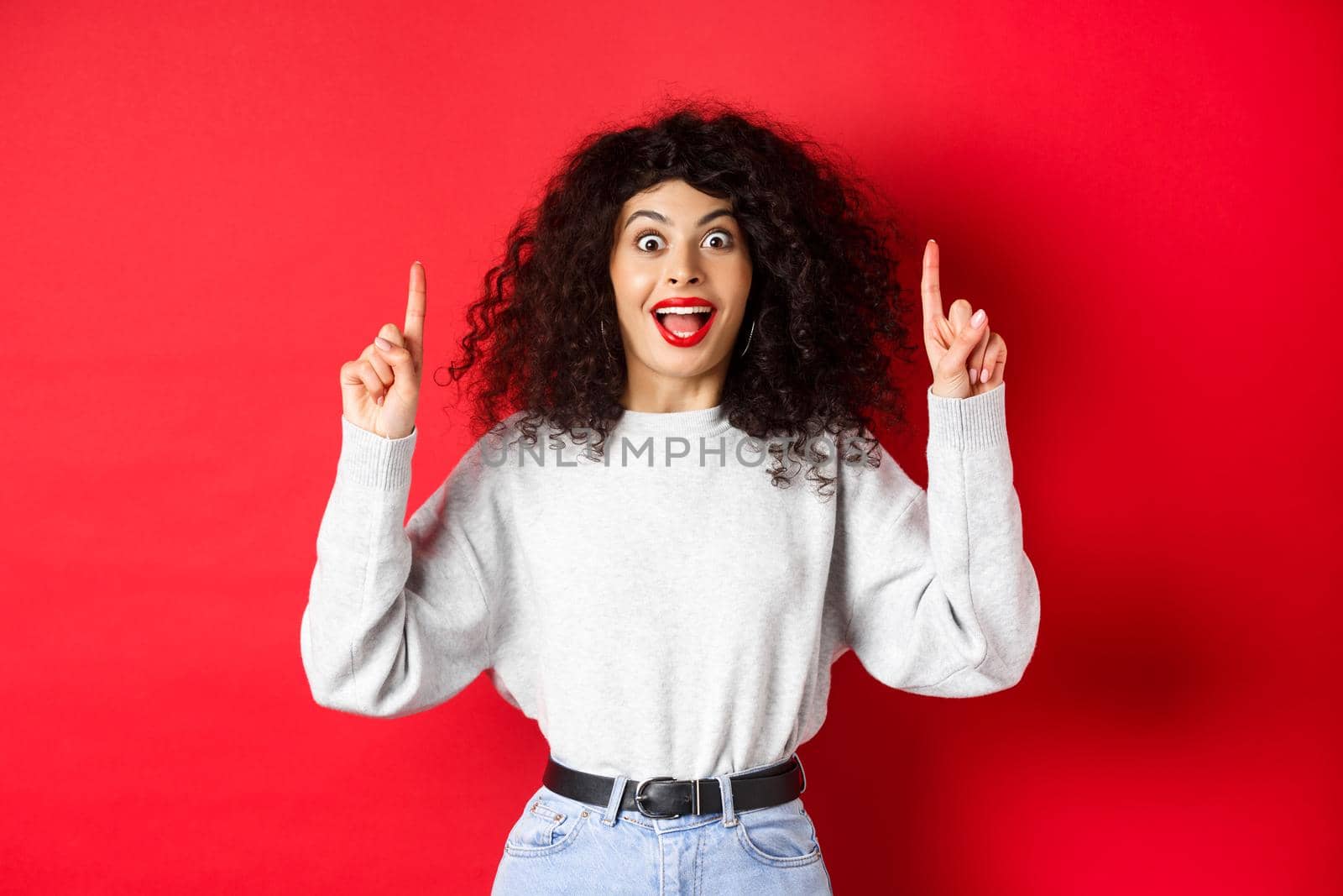 Excited modern girl showing promo, pointing fingers up and smiling amazed, telling big news, standing on red background.