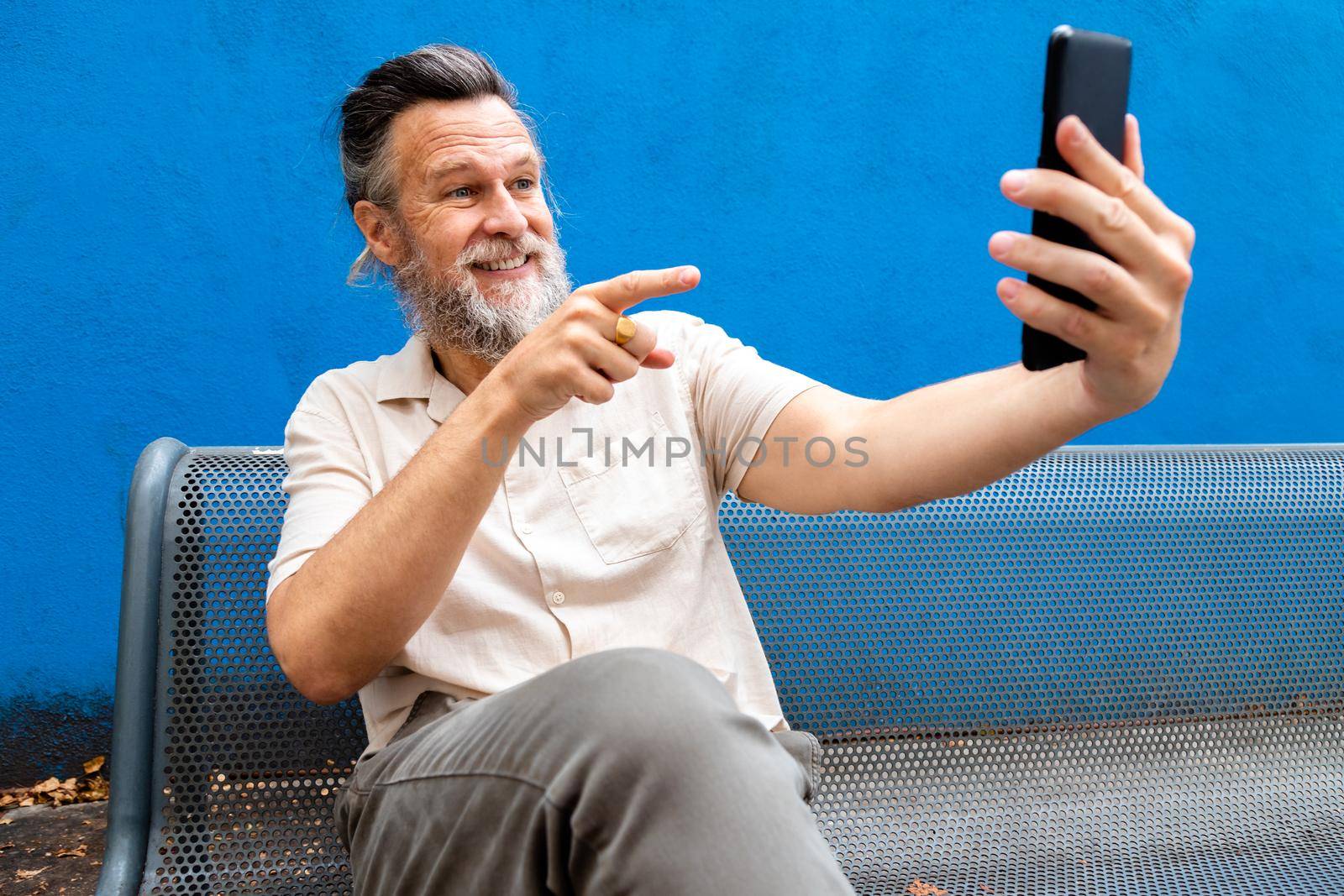 Caucasian mature man with beard sitting on a bench pointing finger to screen during video call. Blue background. Lifestyle concept. Technology concept.