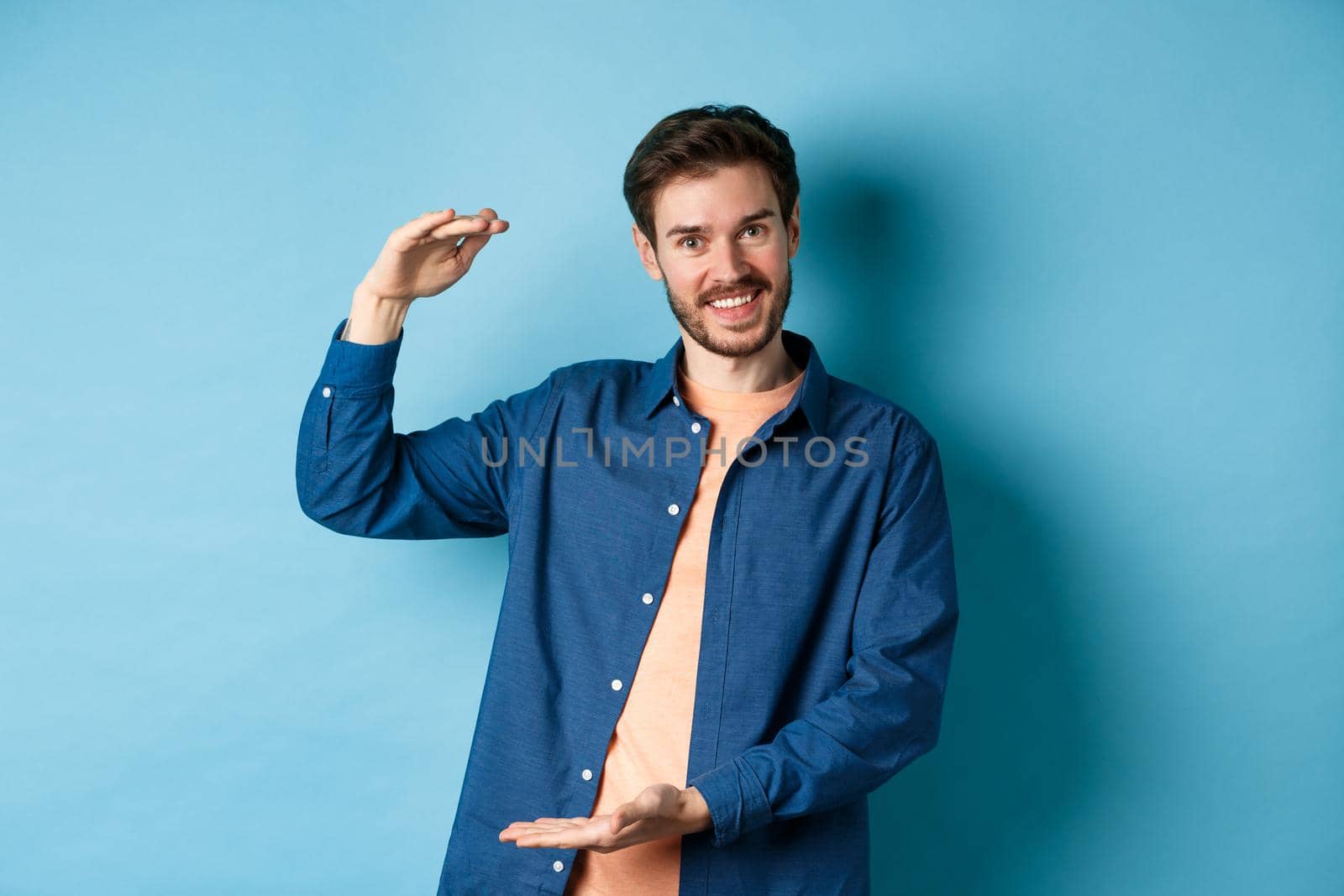 Handsome caucasian guy showing something big, shaping large size object with hands and smiling, standing on blue background.