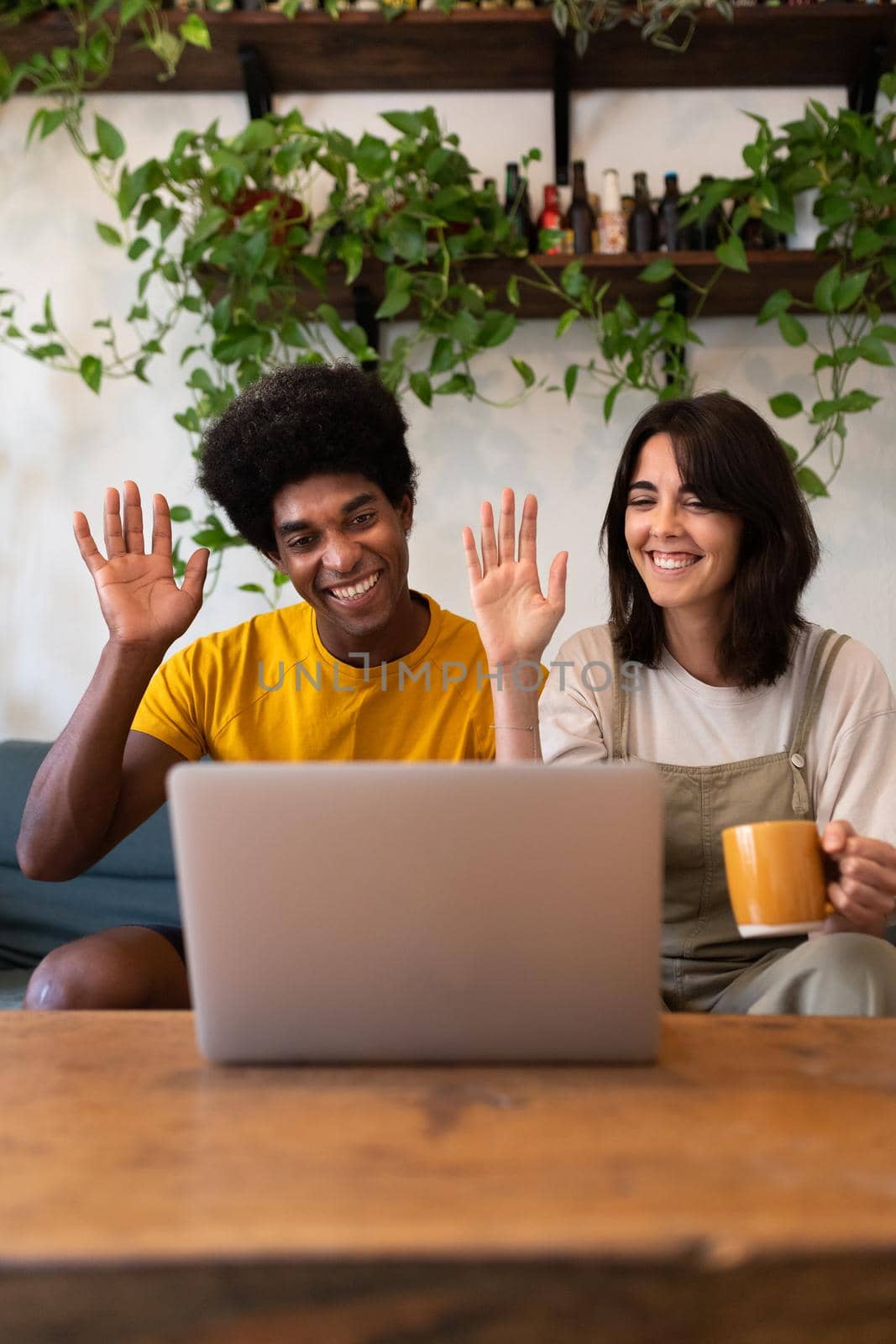 Multiracial couple waves hello to friends on video call using laptop. Vertical. by Hoverstock
