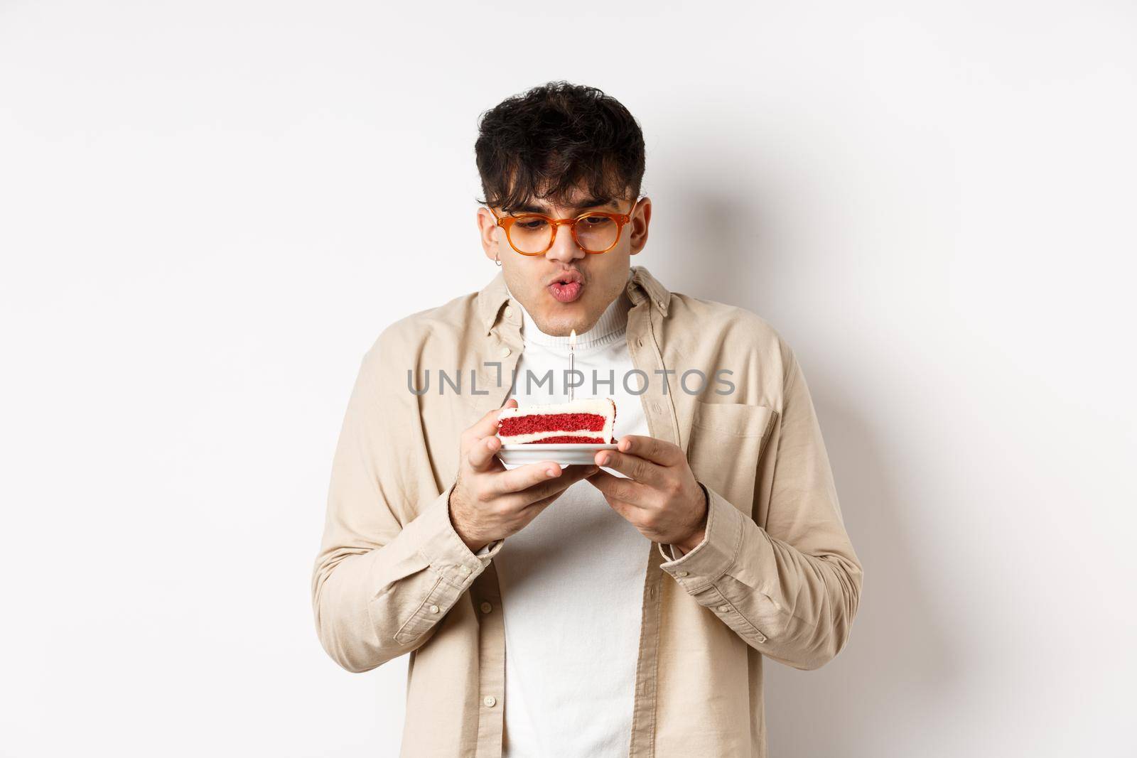 Funny young guy in glasses blowing candle and making wish on birthday cake, standing cheerful against white background.