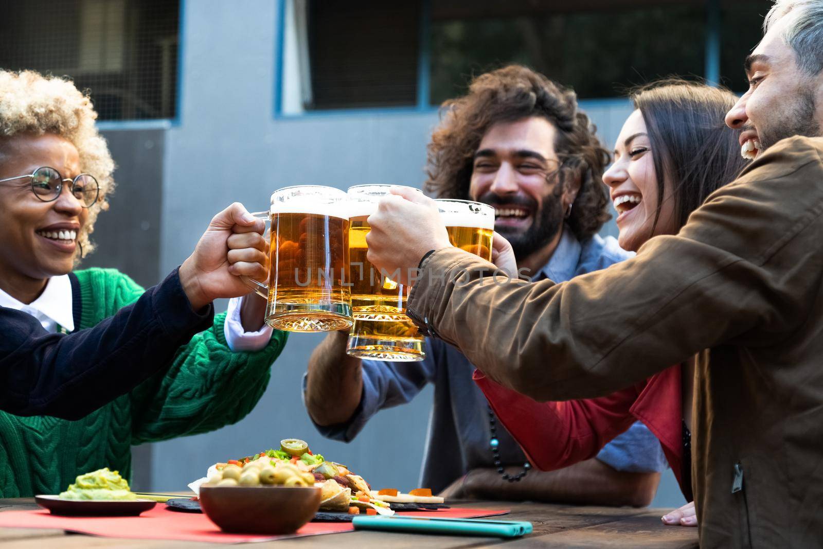 Happy young multiracial friends celebrate toasting with beer in an outdoors bar. by Hoverstock