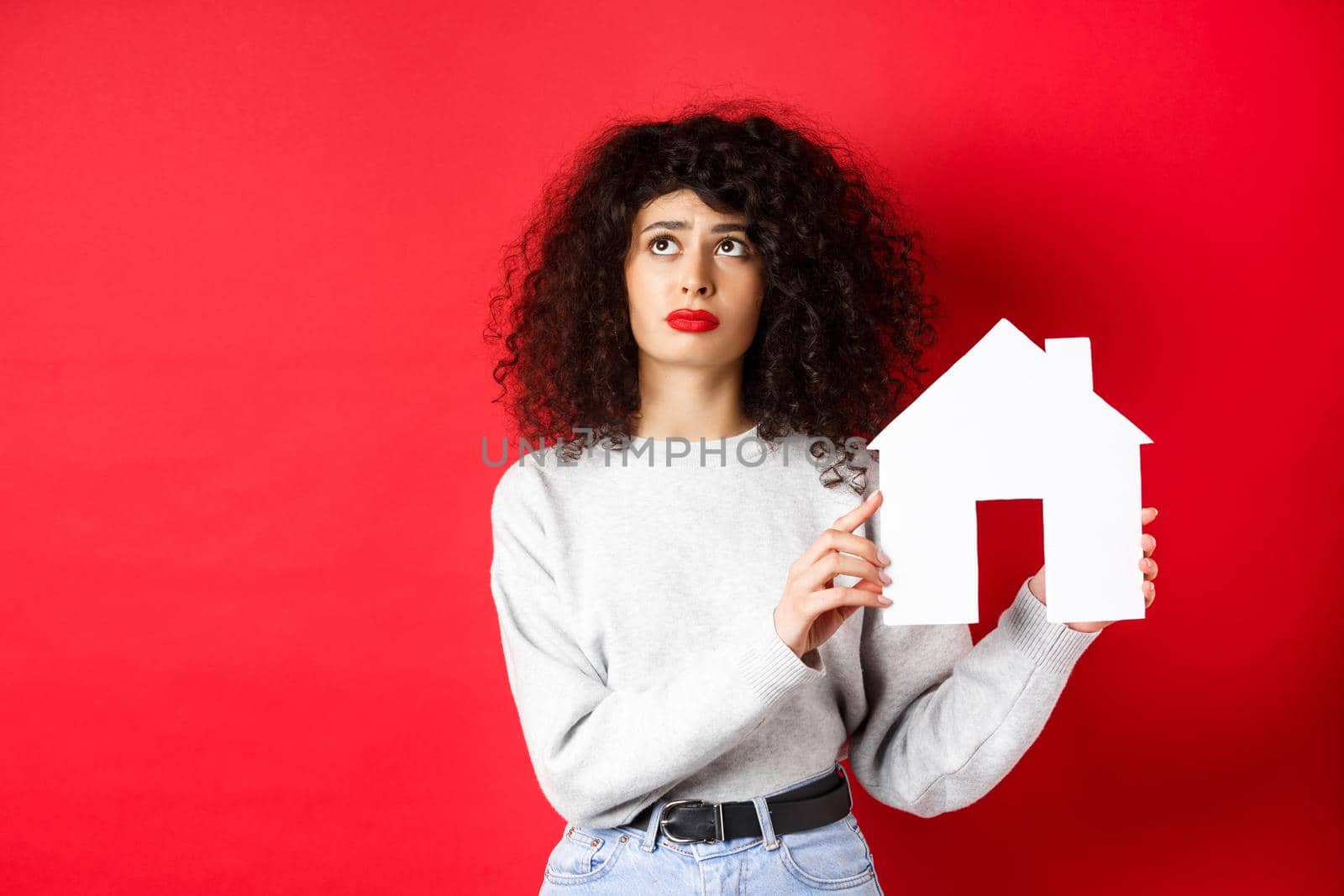 Real estate. Sad woman thinking of buying own house, showing paper home cutout and looking up with uspet face, standing on red background.