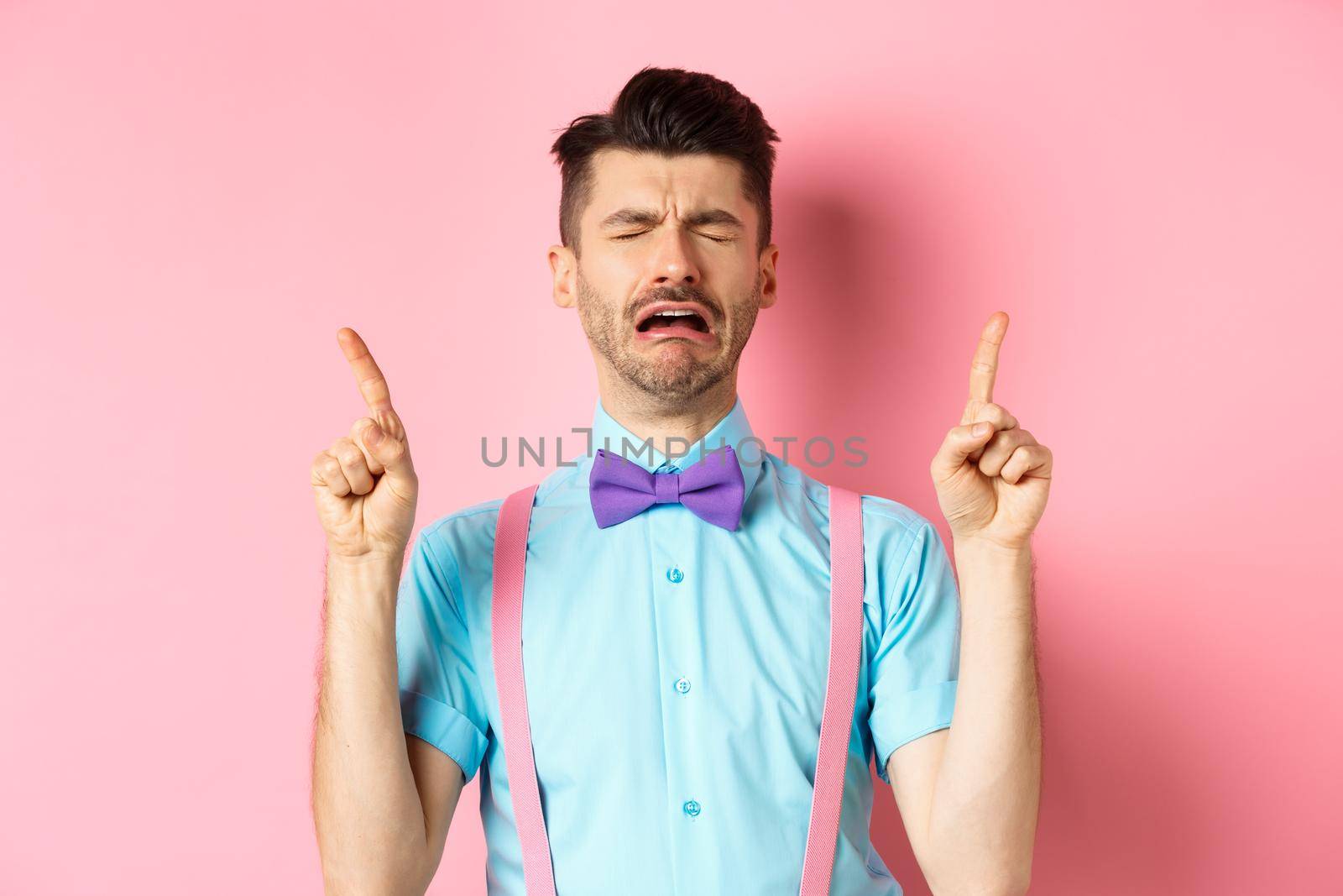 Sad and miserable guy sobbing and crying, pointing fingers up and something disappointed, standing upset on pink background.