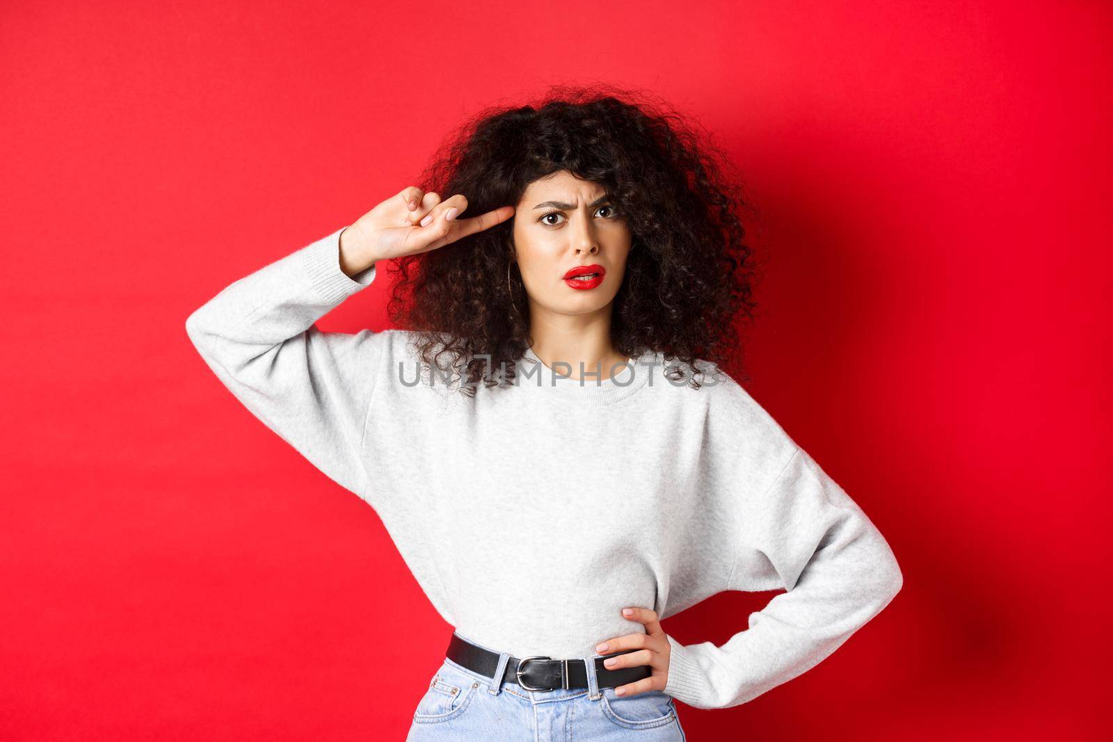 Are you stupid. Annoyed and confused girl with curly hair, pointing at head and scolding person being crazy or strange, standing on red background.