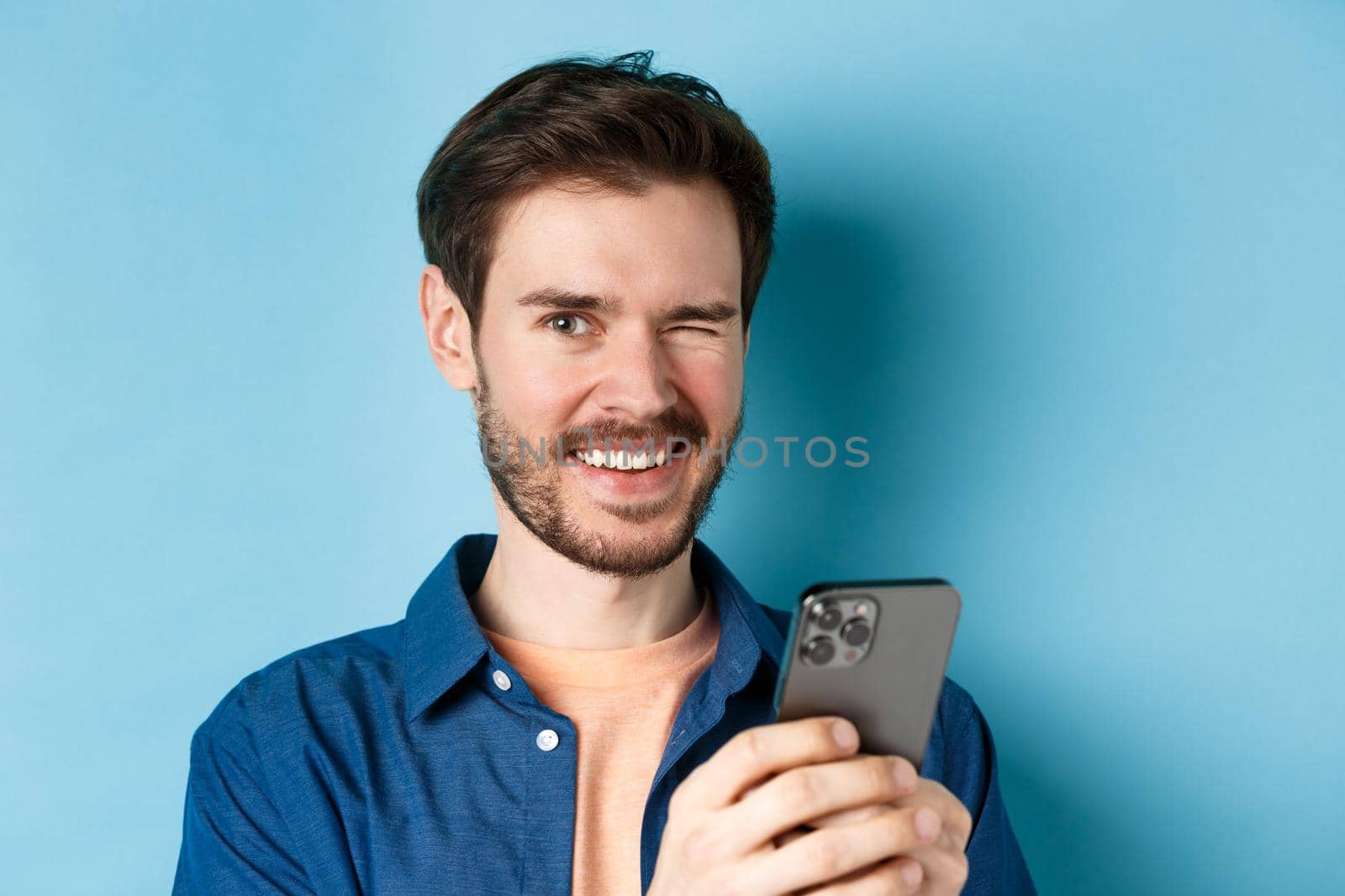 Close up of cheerful man winking and smiling, using mobile phone on blue background.