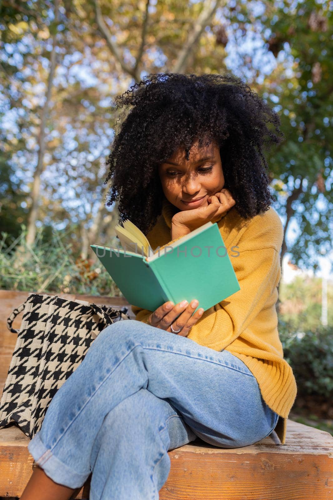 Young African American woman reading a book sitting on park bench. Vertical by Hoverstock