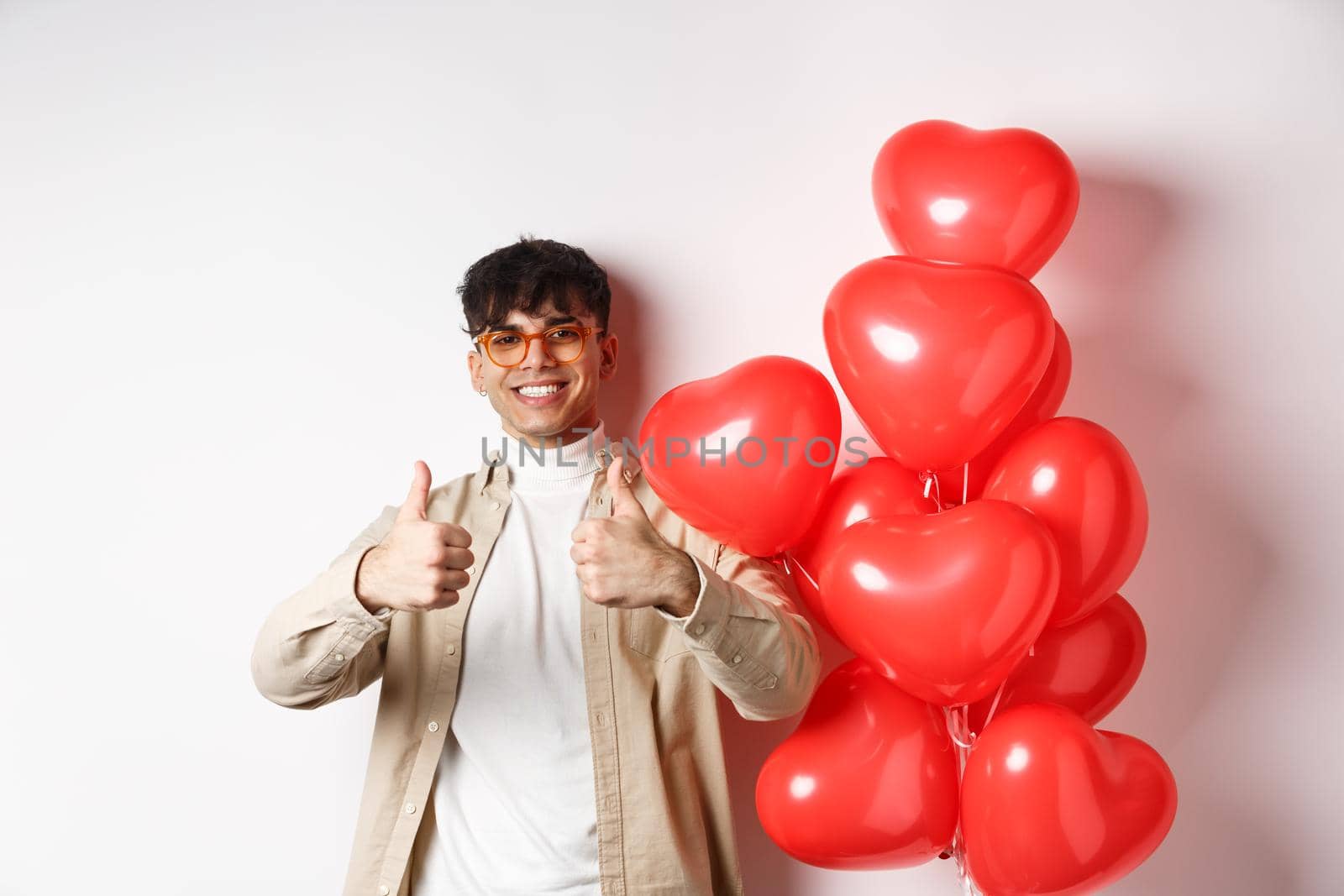 Valentines day and romance concept. Stylish modern man in sunglasses showing thumbs up while standing near heart balloons, saying yes, enjoy date with lover.
