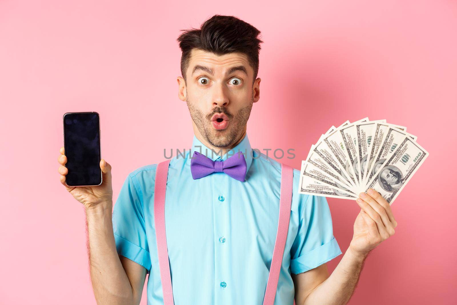 E-commerce and shopping concept. Surprised man showing blank smartphone screen and money, saying wow with amazed face, checking out online offer, pink background.
