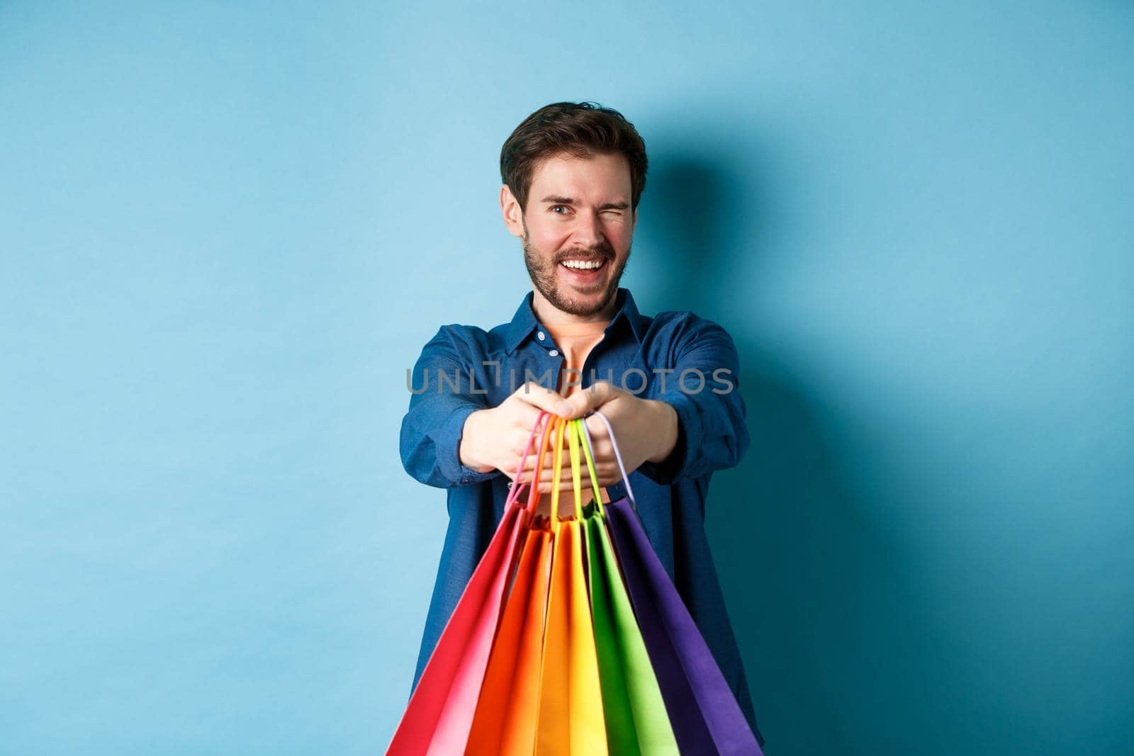 Cheerful handsome man winking and smiling, extending hands with colorful shopping bags, standing on blue background.