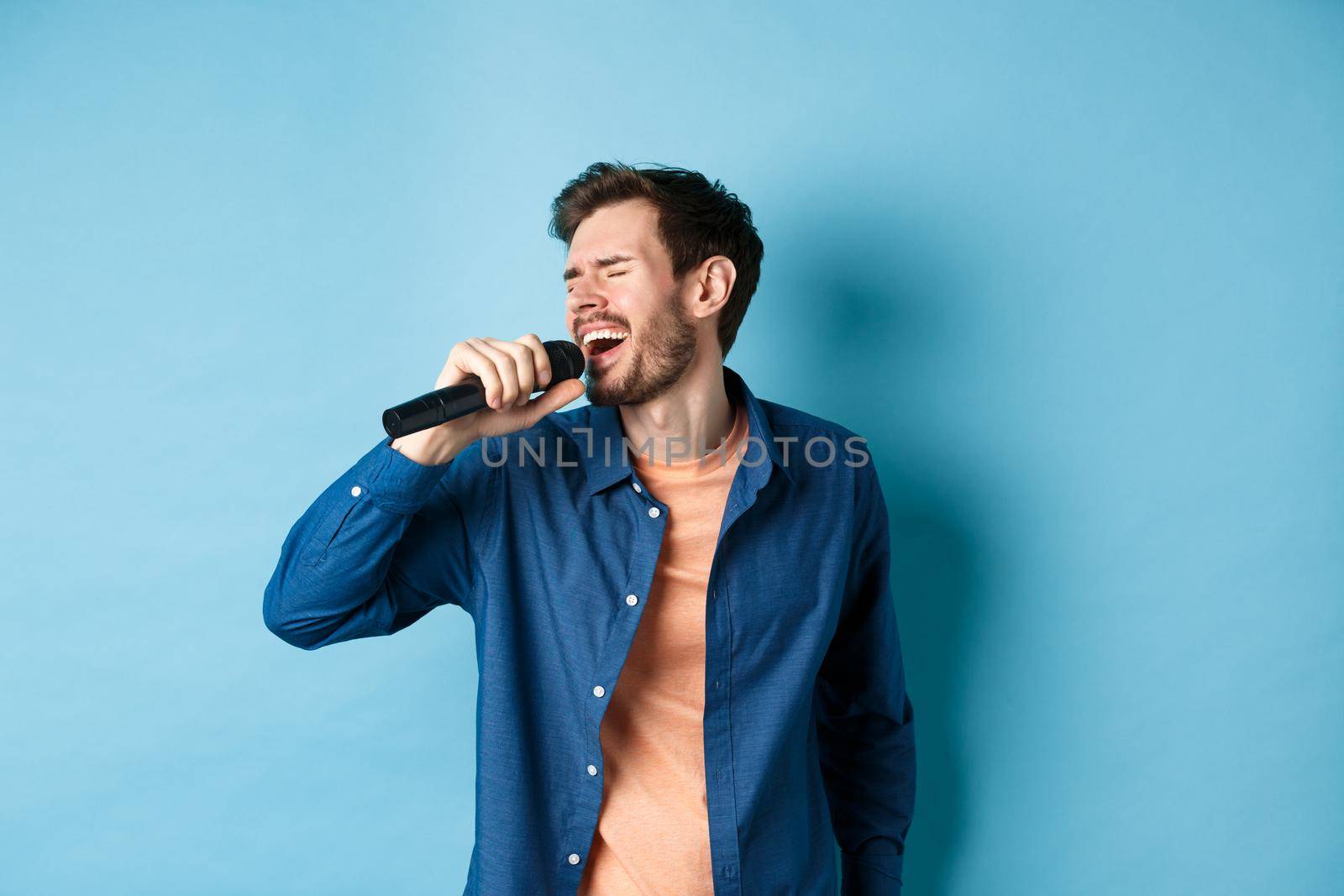 Carefree guy singing karaoke with microphone, perform a song, standing on blue background. Copy space