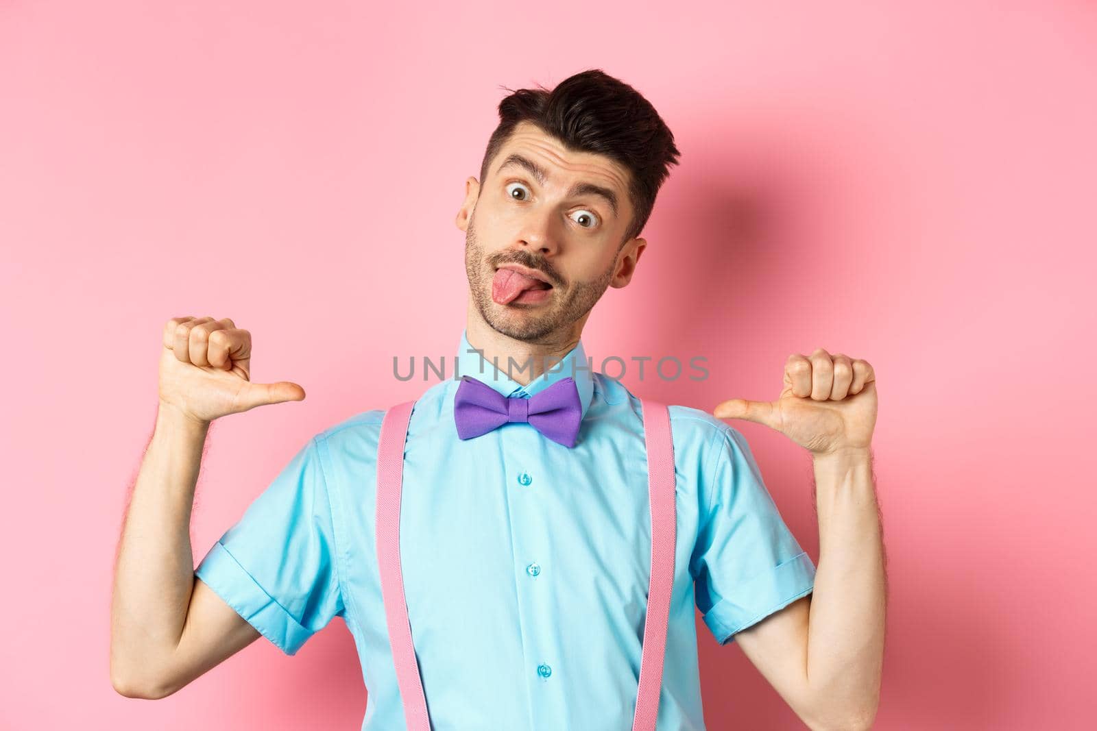 Funny guy in suspenders and bow-tie showing tongue, pointing at himself as if self-promoting, standing over pink background.