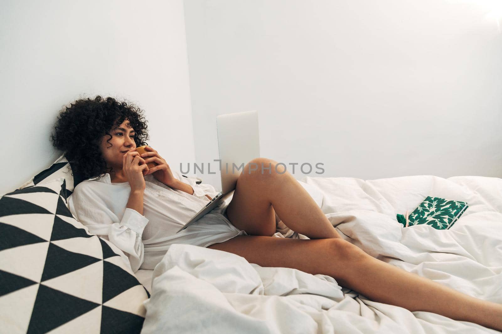 Young mixed race woman lying on bed using laptop and drinking coffee. Copy space. Lifestyle and technology concept.