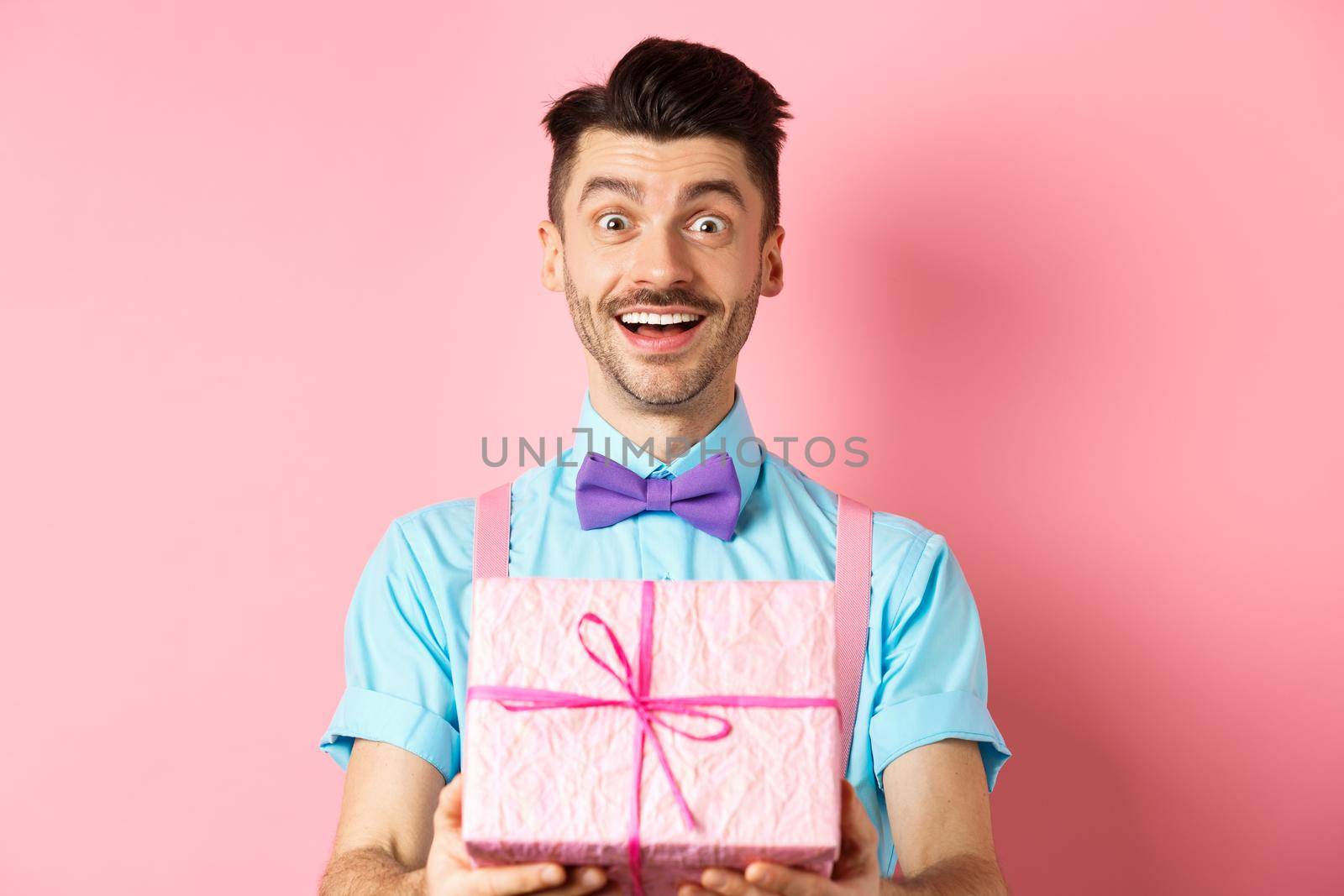 Holidays and celebration concept. Cheerful guy wishing happy birthday and giving you gift wrapped in box, standing over pink background in festive clothes.