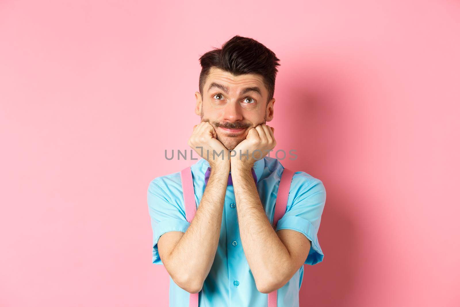 Silly young man leaning on hands and looking up, dreaming of something, imaging things on pink background.