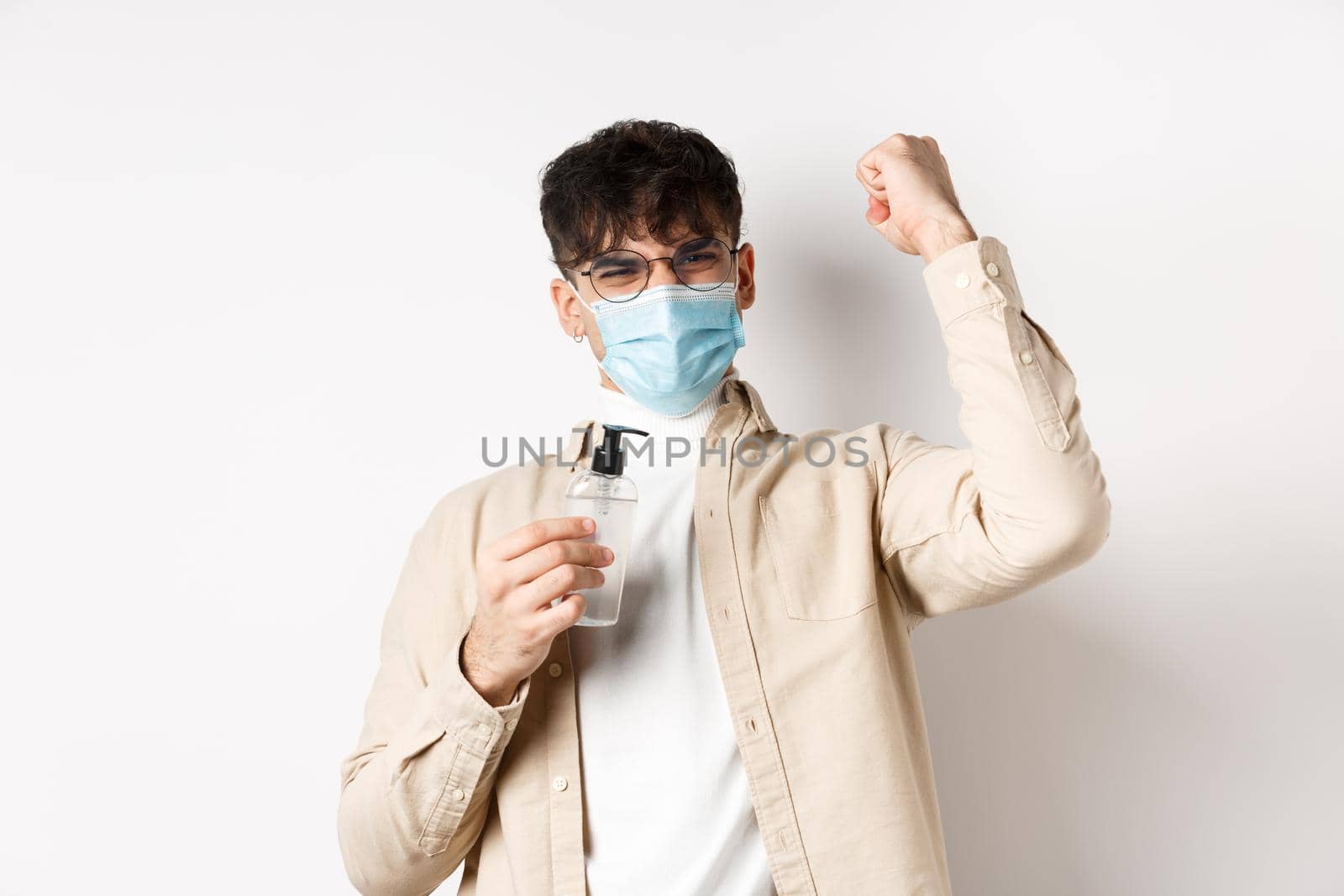 Health, covid and quarantine concept. Happy young man motivated to fight coronavirus, showing hand sanitizer and raising hand up in triumph, standing on white background.
