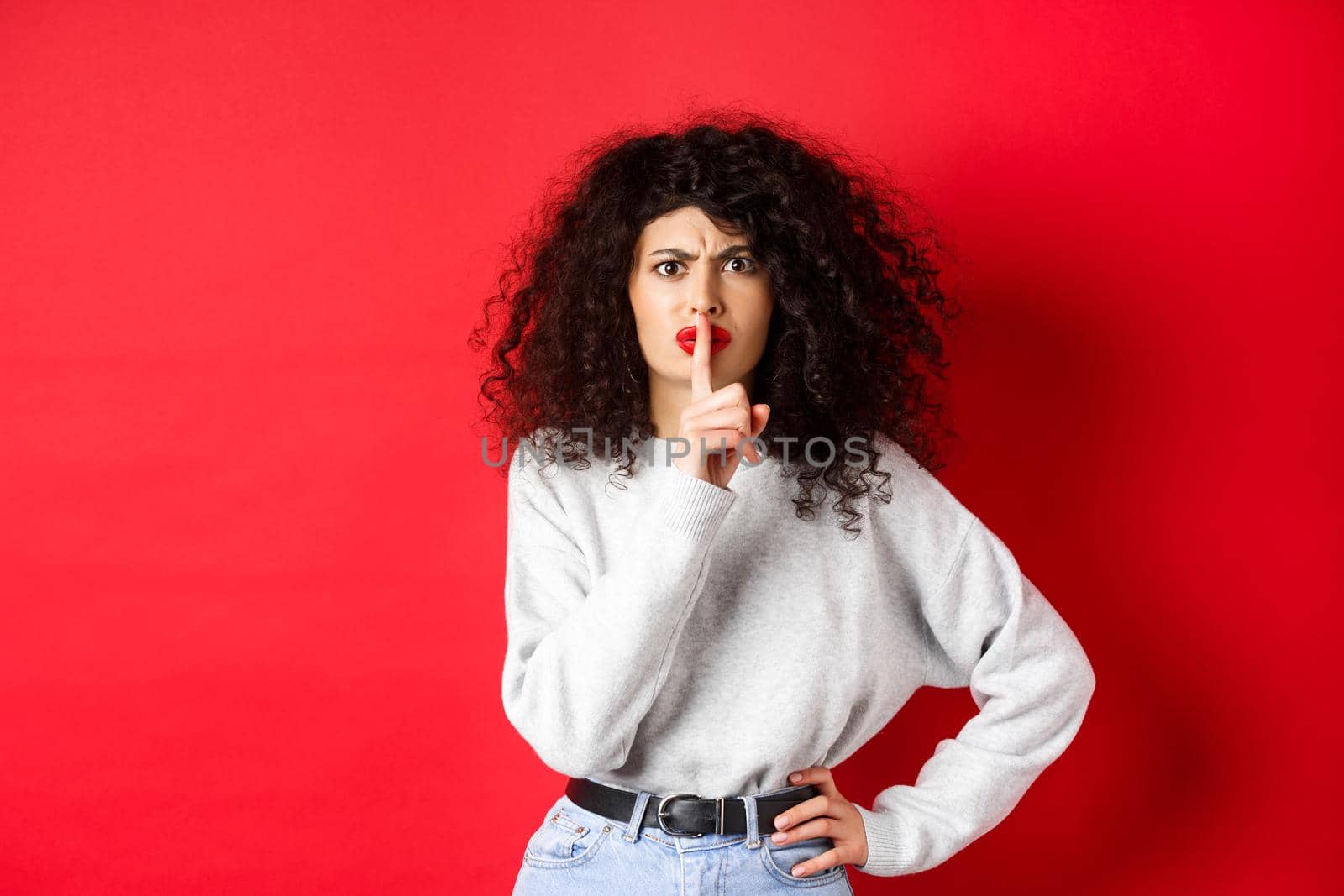 Angry young woman with curly hair and red lips, frowning and shushing, tell to be quiet, make taboo gesture to keep mouth shut, standing on red background.