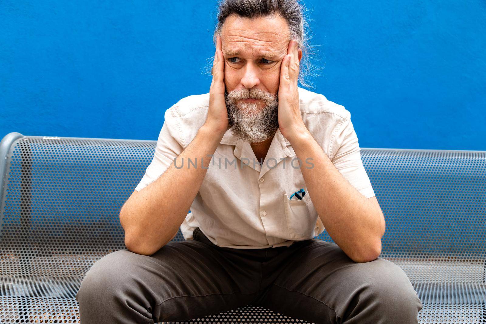Pensive and worried caucasian mature man with hands on face sitting on a bench.Exhausted expression.