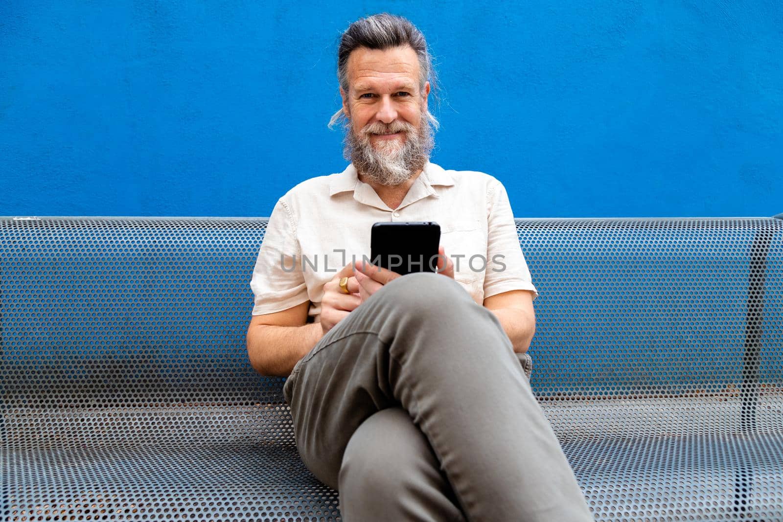 Mature caucasian man with beard sitting on metal bench using phone looking at camera. Lifestyle concept.