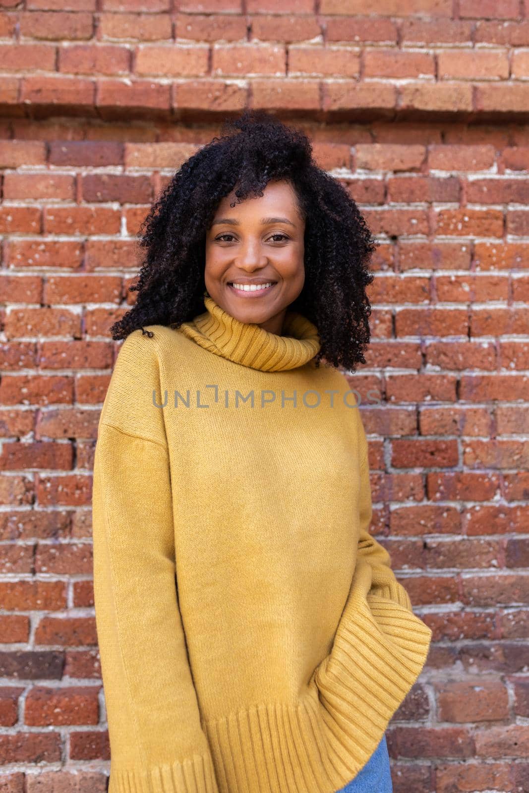 Vertical portrait of African American woman with curly hair wearing yellow sweater. Brick background wall. by Hoverstock