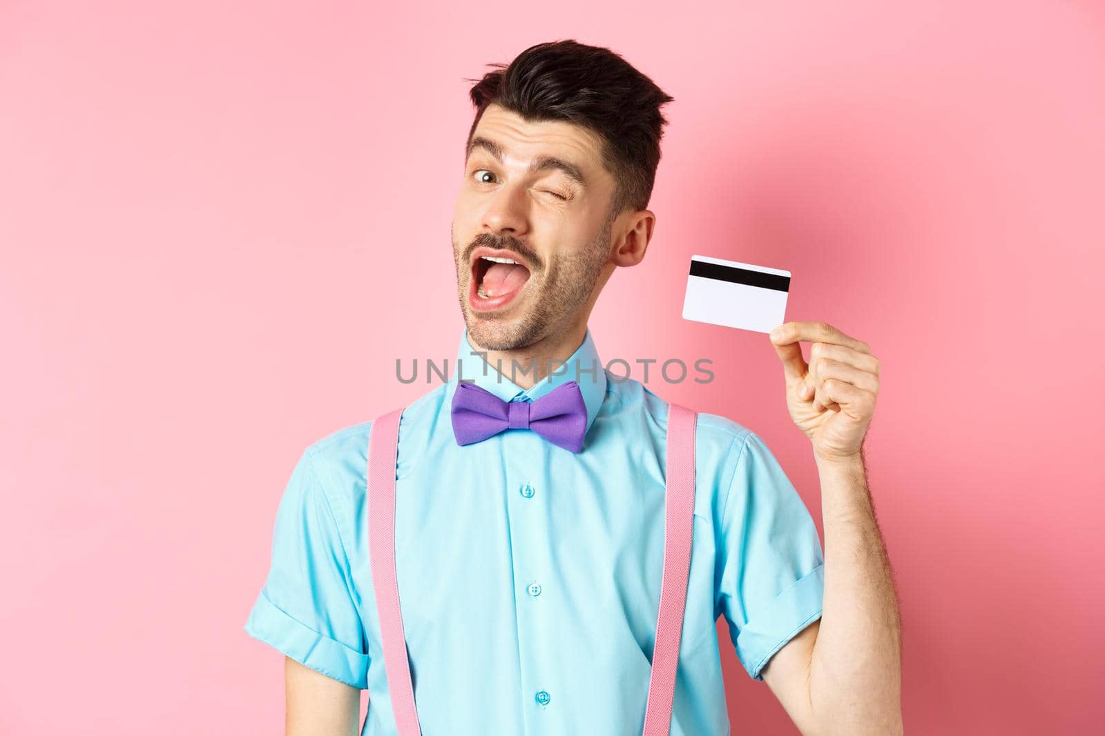 Shopping concept. Funny guy with moustache winking at camera, showing plastic credit card, recommending bank promo offer, standing on pink background.