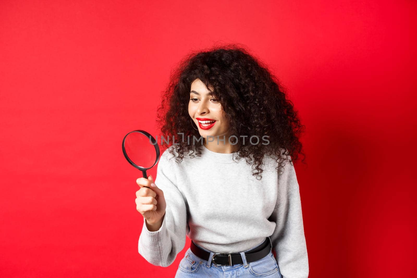 Young woman looking through magnifying glass, investigating or searching for something, stare curious aside, found interesting thing, standing on red background.