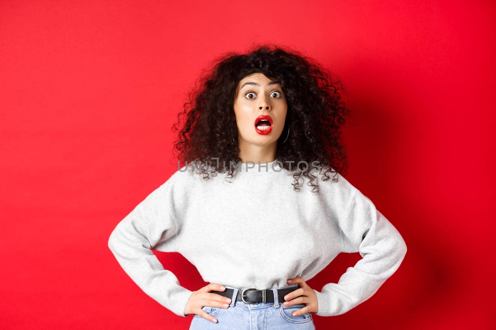 Shocked italian woman with curly hair, gasping and staring at camera astonished, open mouth, standing in white sweatshirt on red background.