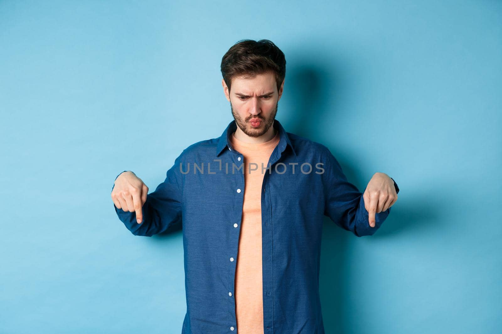 Confused and shocked guy looking and pointing down at something strange, standing on blue background. Copy space