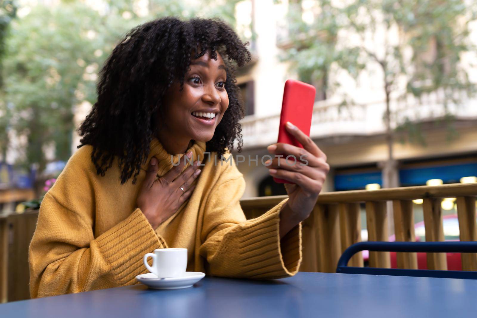 Young African American woman with surprise expression on video call using smartphone in outdoors coffee shop. Copy space. Lifestyle and technology concept.
