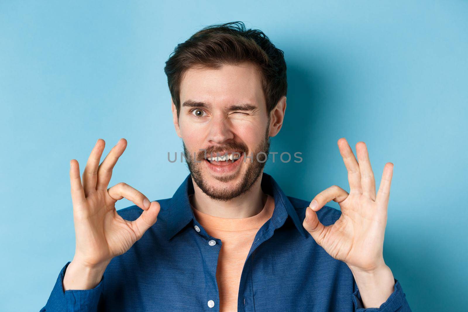 Close up of handsome confident guy winking, showing okay gestures, guarantee quality, recommending something good, standing on blue background.