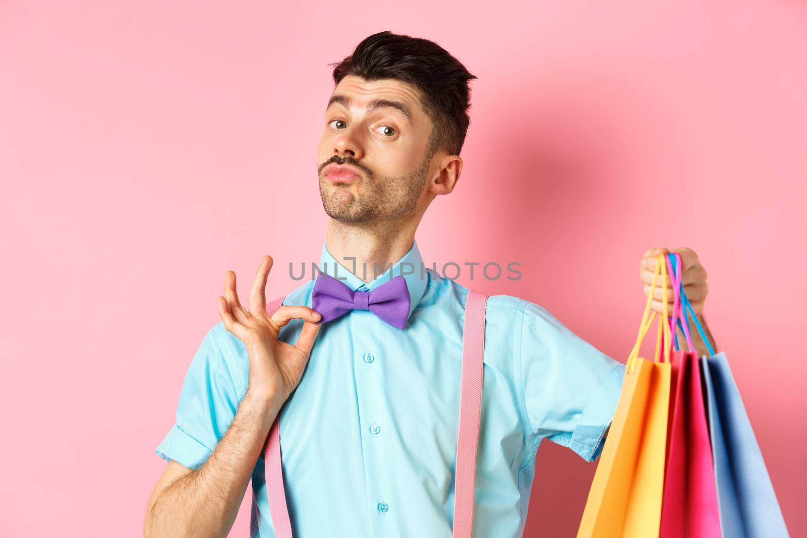 Fancy guy with moustache fixing his bow-tie and holding shopping bags, boyfriend bring gift packages, standing over pink background by Benzoix