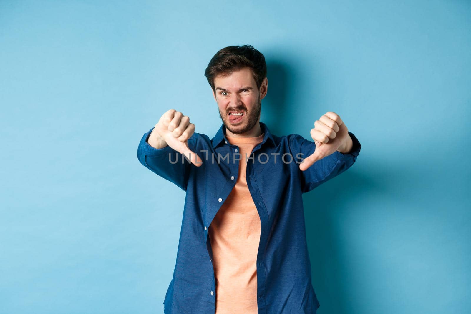 Disgusted guy express negative opinion, showing thumbs down and tongue, frowning upset, disapprove something bad, standing on blue background.