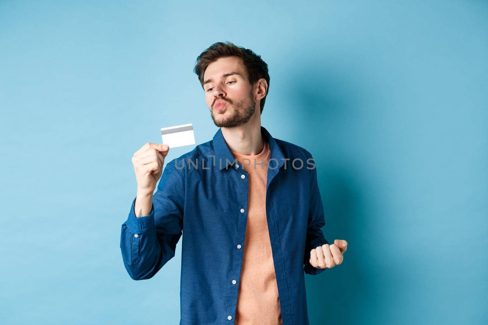 Funny guy kissing his plastic credit card and looking satisfied, shopping with discounts, standing on blue background by Benzoix