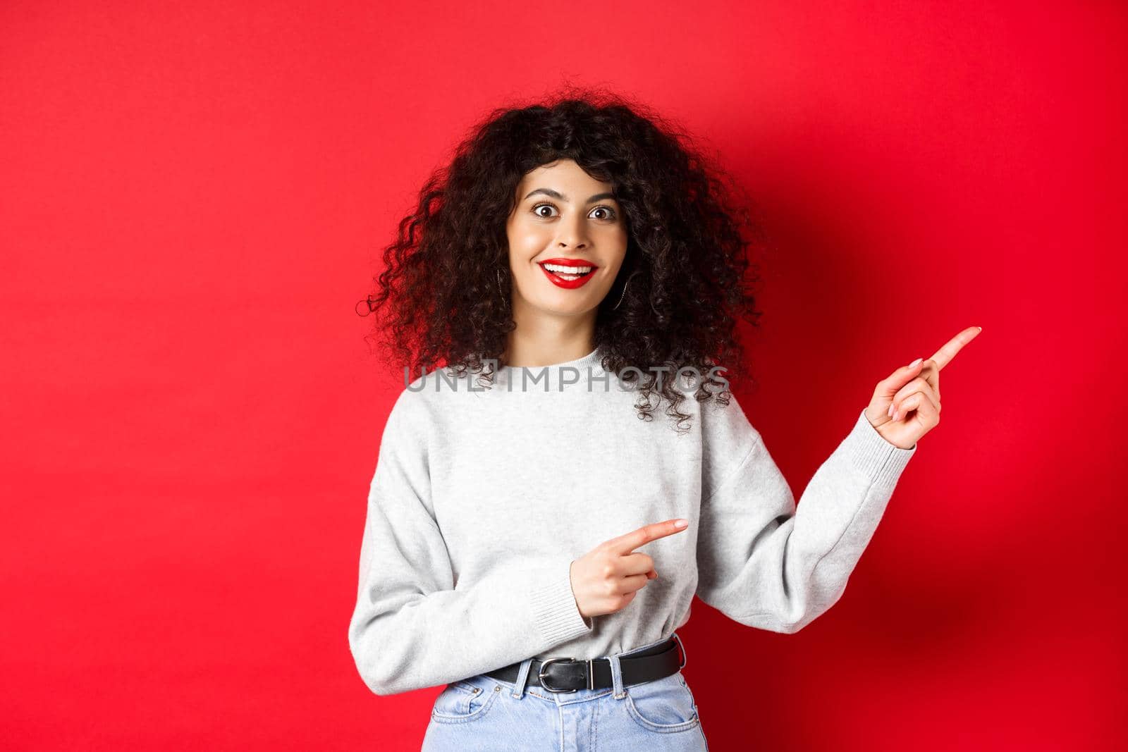 Amused young woman with curly hairstyle, pointing fingers right at logo, looking impressed and excited, checking out promotion, red background.