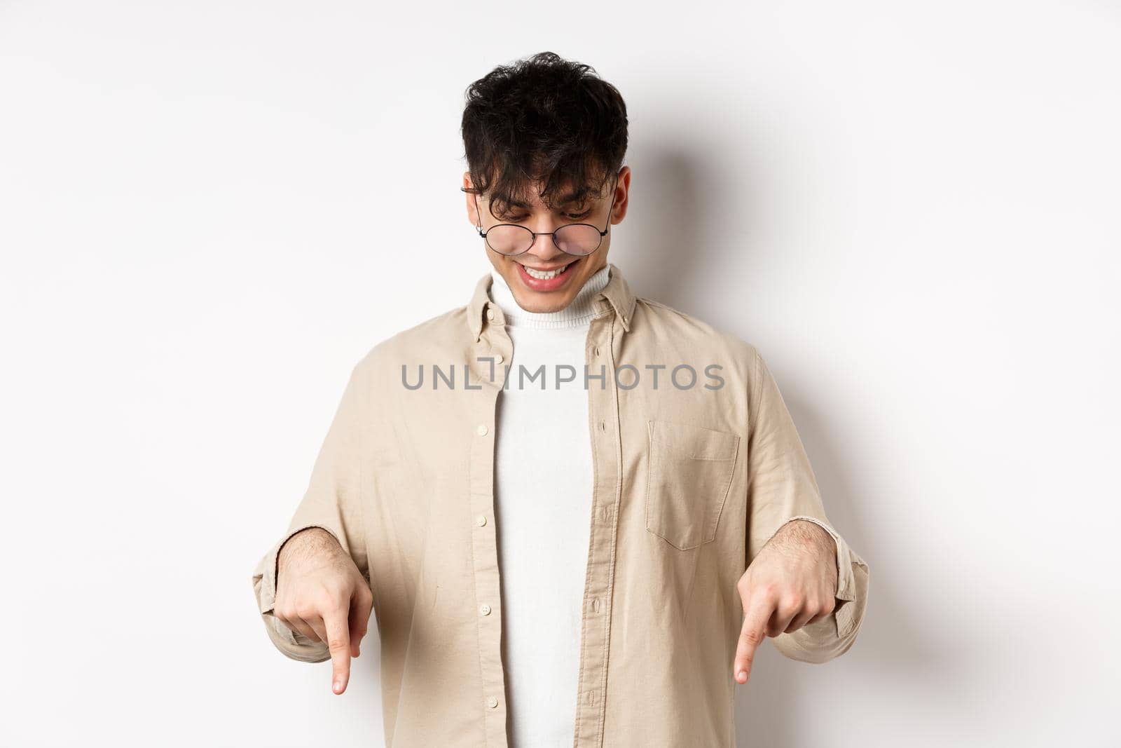 Stylish modern guy in glasses pointing, looking down with happy smile, showing banner, standing on white background.