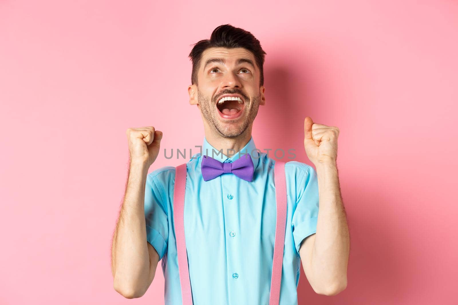 Relieved and happy man thanking god, looking up in sky while celebrating victory, winning prize and cheering, triumphing over pink background by Benzoix