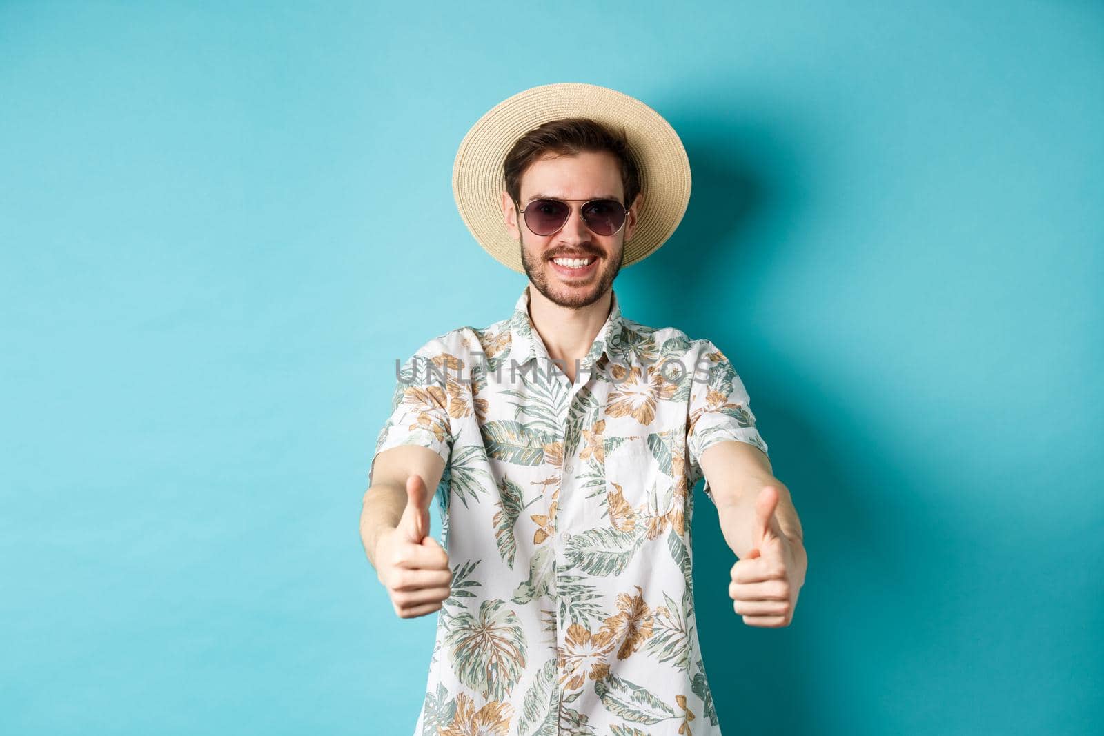 Happy tourist in sunglasses and straw hat showing thumbs up and smiling, enjoying summer holiday, standing on white background.