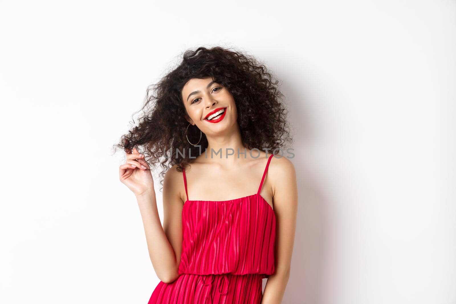 Beautiful woman with curly hair, wearing red dress and lipstick, playing with curl strand and smiling at camera, white background by Benzoix