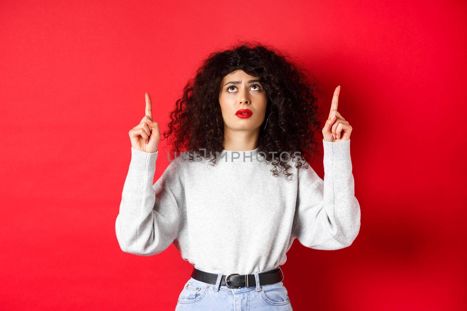 Concerned young woman with curly hair, frowning and looking doubtful, pointing up with hesitant or worried face, standing on red background.