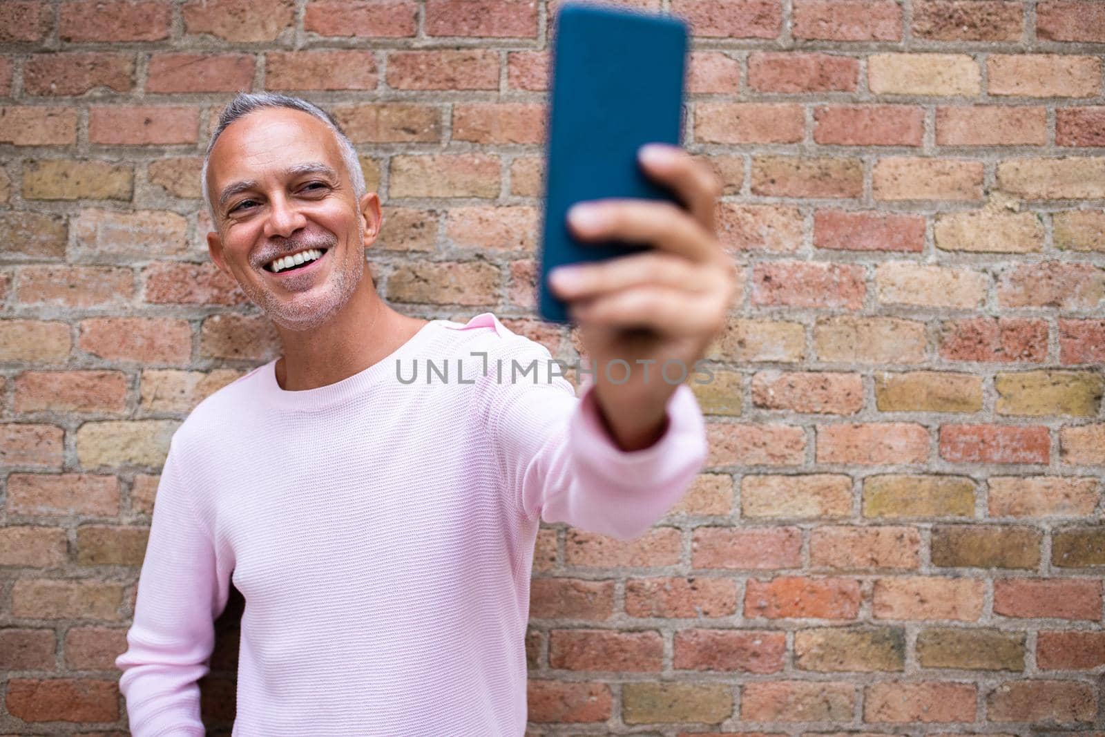 Smiling caucasian adult man taking selfie. Orange brick wall background. Copy space. Technology and lifestyle concept.