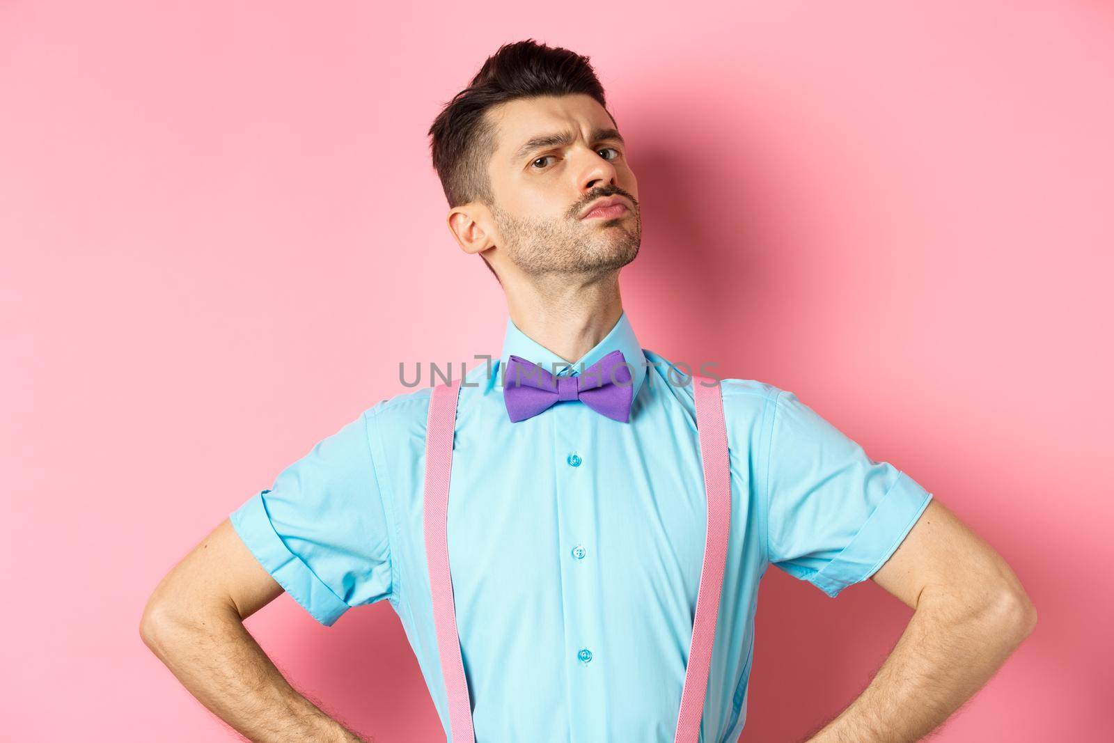 Proud and confident caucasian guy with moustache and bow-tie, looking arrogant with chin up, frowning and looking at camera, standing on pink background.