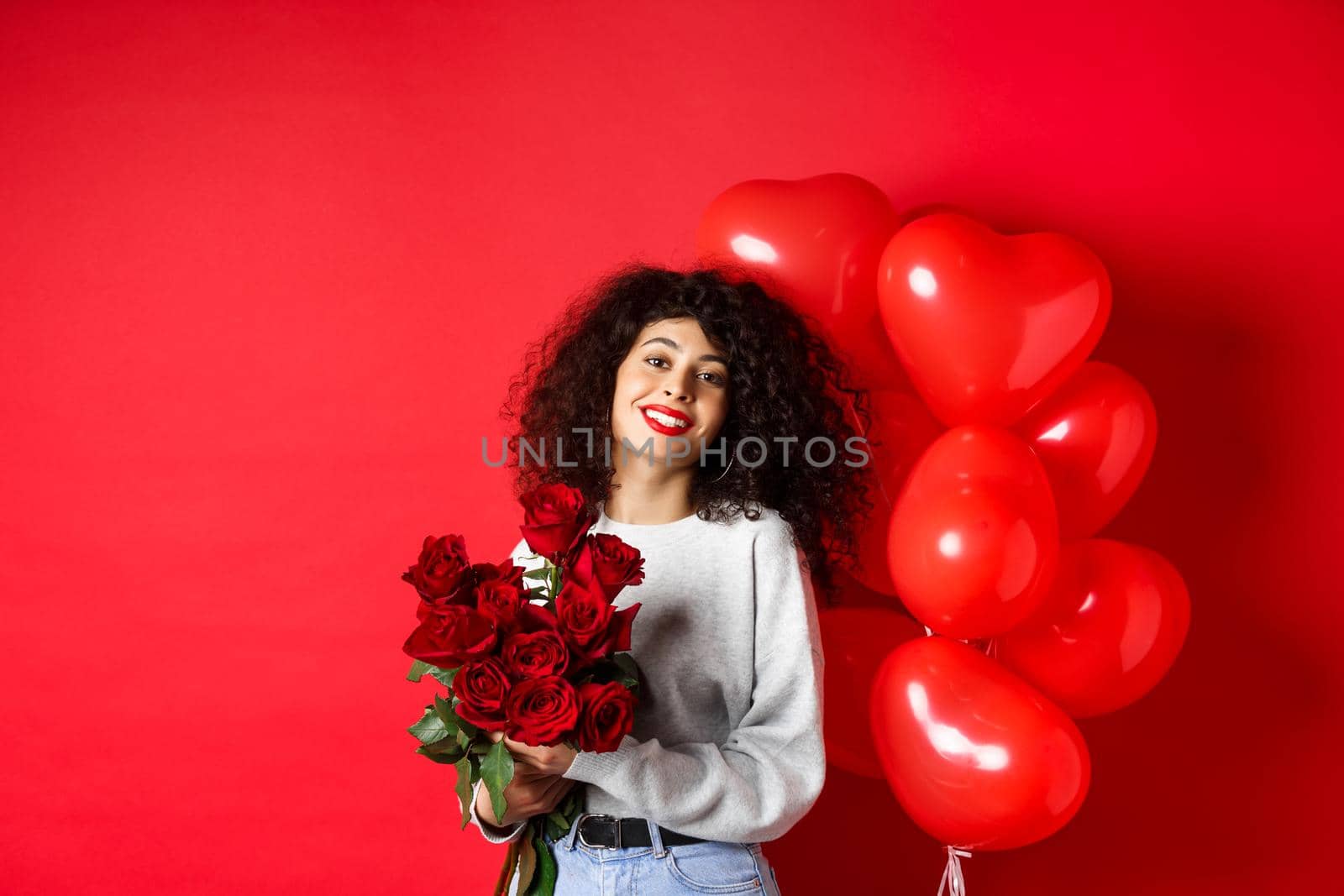 Holidays and celebration. Happy beautiful woman with curly hair, receive bouquet of roses and smiling, standing near party balloons, red background.