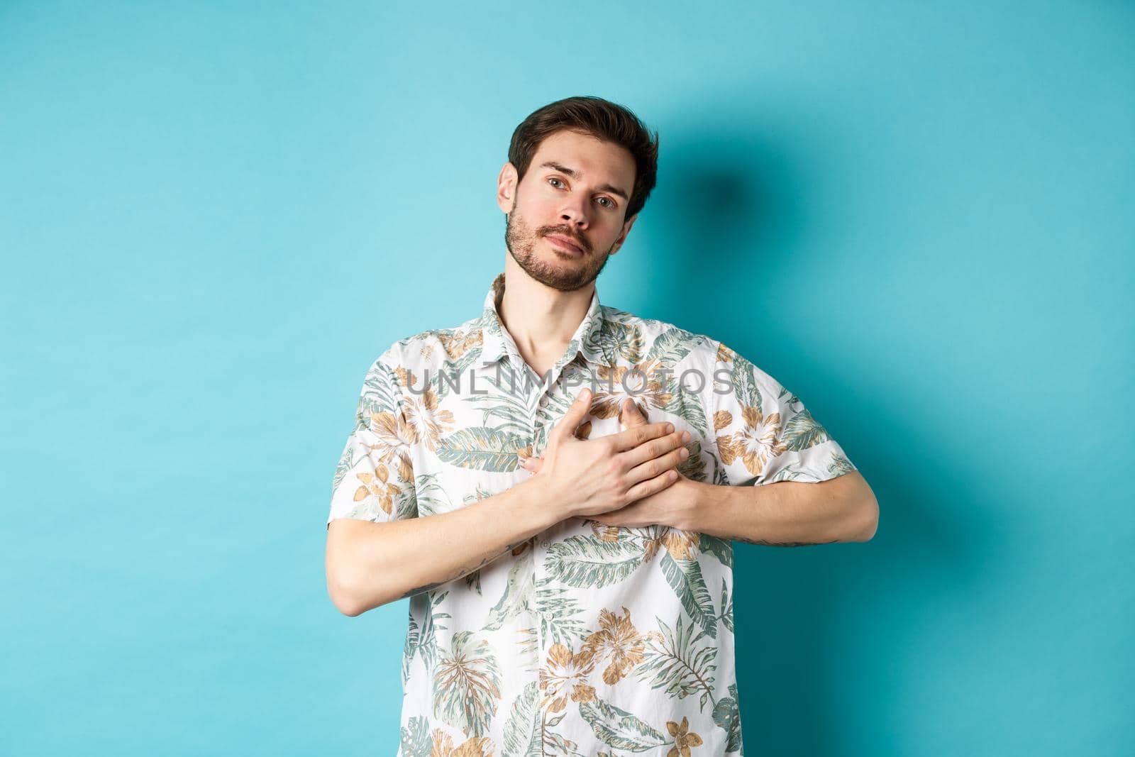 Handsome young man in summer shirt, holding hands on heart and looking thankful, being grateful, standing on blue background by Benzoix