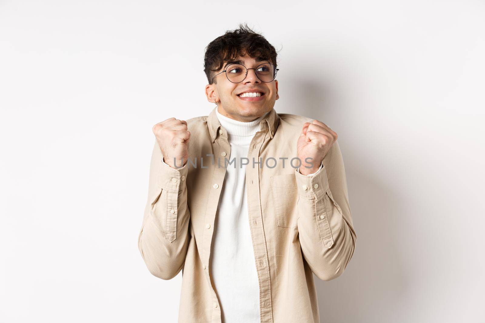 Image of handsome excited man feeling motivated and lucky, looking right and smiling, making fist pump gesture to celebrate victory, winning prize, standing on white background.