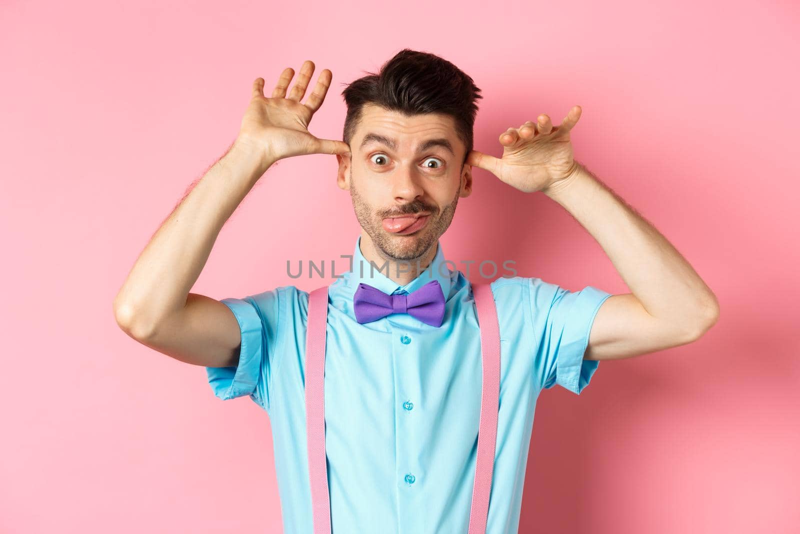 Silly and childish guy in suspenders and bow-tie, mocking someone, fooling around and making funny gestures, standing on pink background.