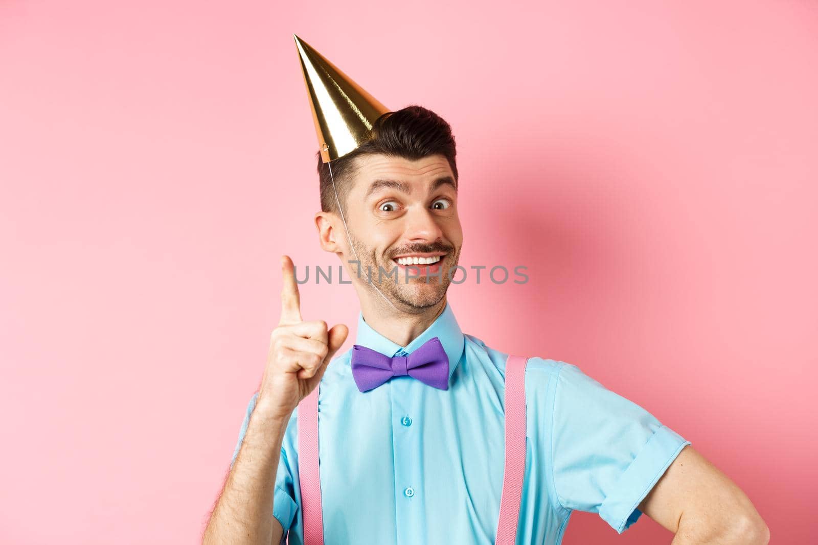 Holidays and celebration concept. Image of happy man in birthday party hat pitching an idea, raising finger and smiling, have plan or solution, standing over pink background.